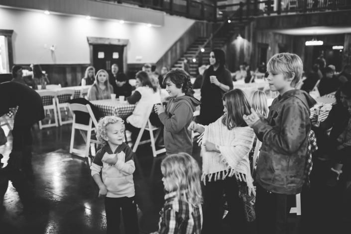 Children gathered in a hall, standing and chatting near tables and stairs at an indoor event.