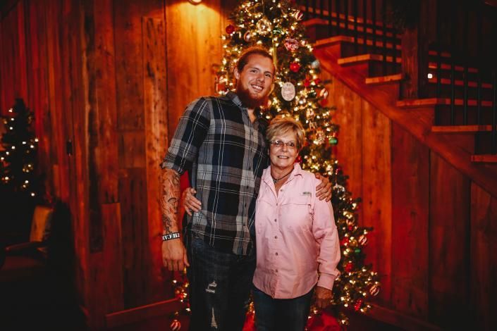 Two people posing by a lit Christmas tree and wooden staircase in a warmly decorated room