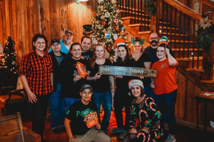 Group of people posing indoors by a Christmas tree, smiling and holding a festive sign.