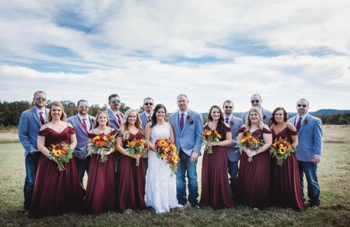 Wedding party posing outdoors in blue sky, with bride, groom, and attendants holding bouquets.