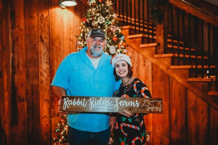 Couple holding a wooden holiday sign in front of a lit Christmas tree on a wood staircase