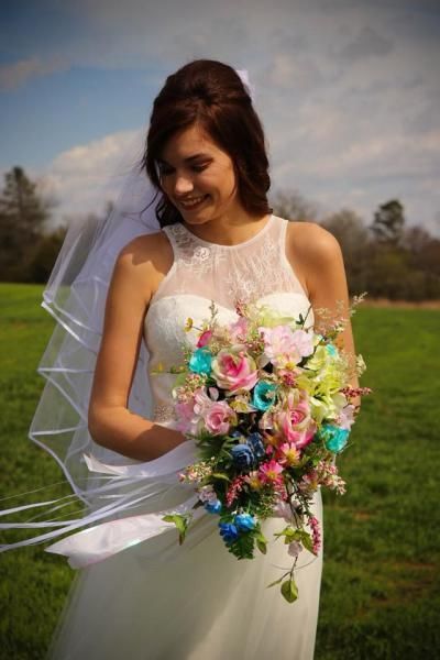Woman in a white dress holding a colorful bouquet outdoors, smiling with her veil blowing in the wind.