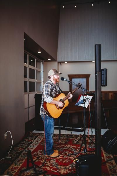 Musician singing and playing acoustic guitar on a small indoor stage with a microphone and music stand