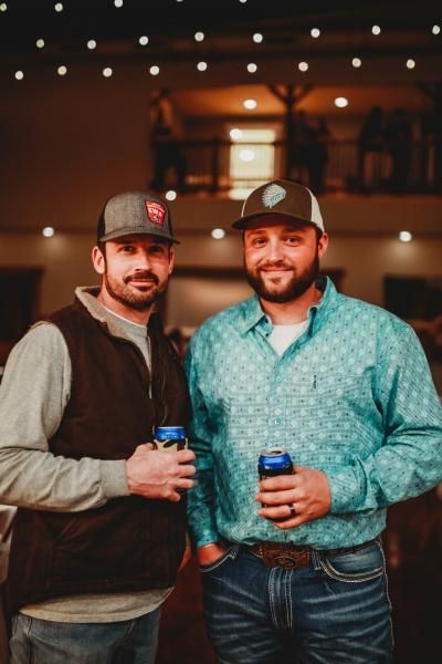 Two men in casual clothes and caps holding drinks, standing indoors under warm string lights.