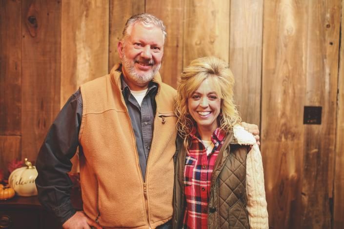 Two smiling people standing indoors against a wooden wall, wearing tan and red plaid jackets.