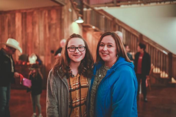 Two people smiling indoors, one in glasses and plaid, beside a staircase in warm lighting
