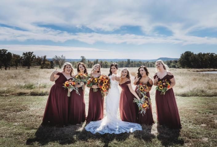 Bridal party in burgundy dresses with bouquets stands outdoors beside the bride in a white gown.