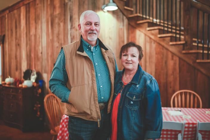 Two people standing in a rustic wooden room, smiling beside a table with a red-checkered cloth