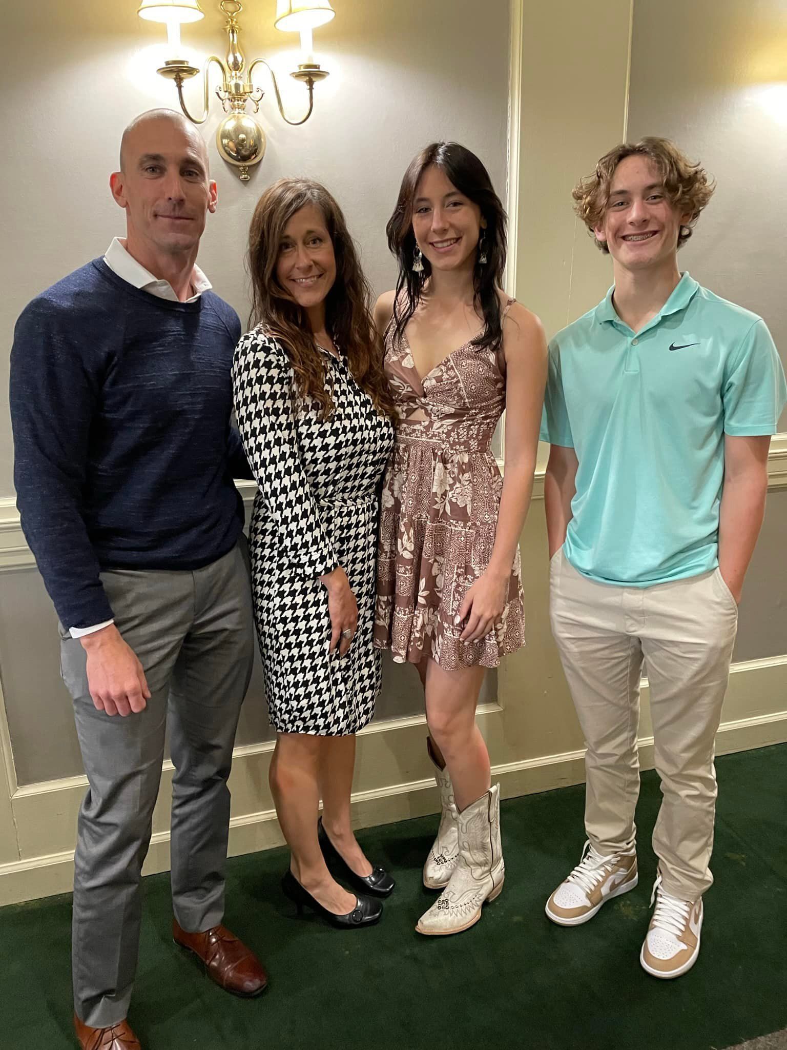 Four people standing together indoors, dressed in semi-formal outfits, smiling beneath a chandelier.