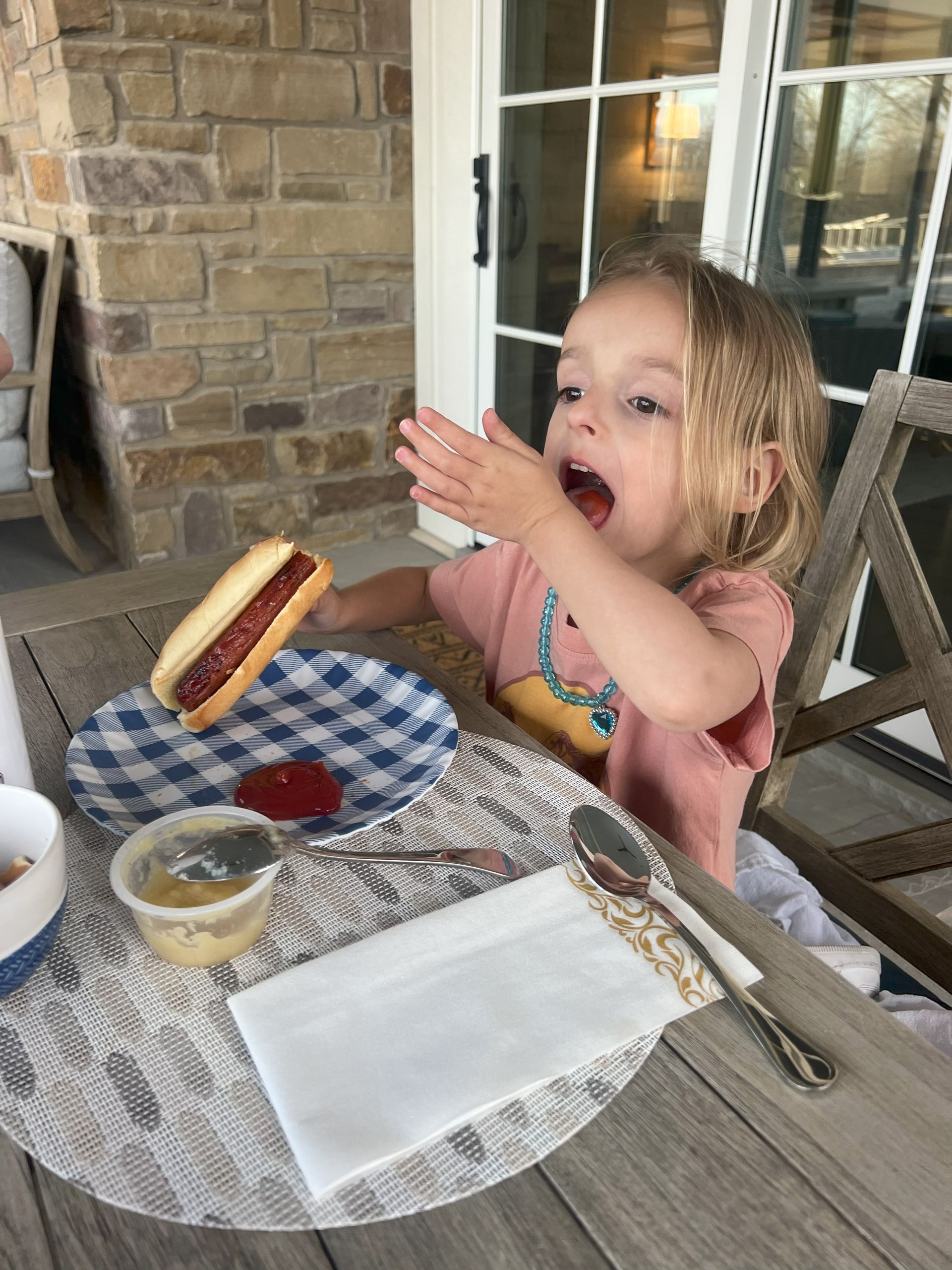 Child at an outdoor table eating a hot dog, with a plate, napkin, and drink on a patio