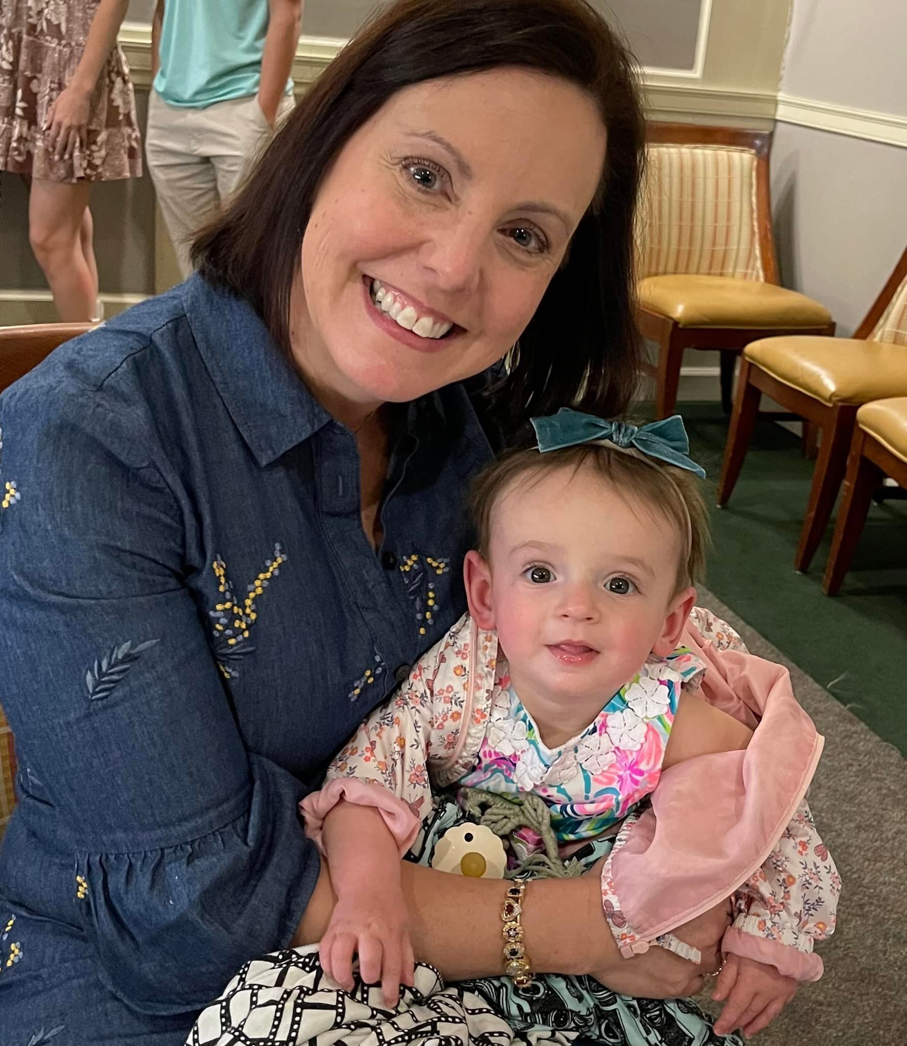 Smiling woman in denim shirt holding a baby with a pink blanket in a cozy indoor setting
