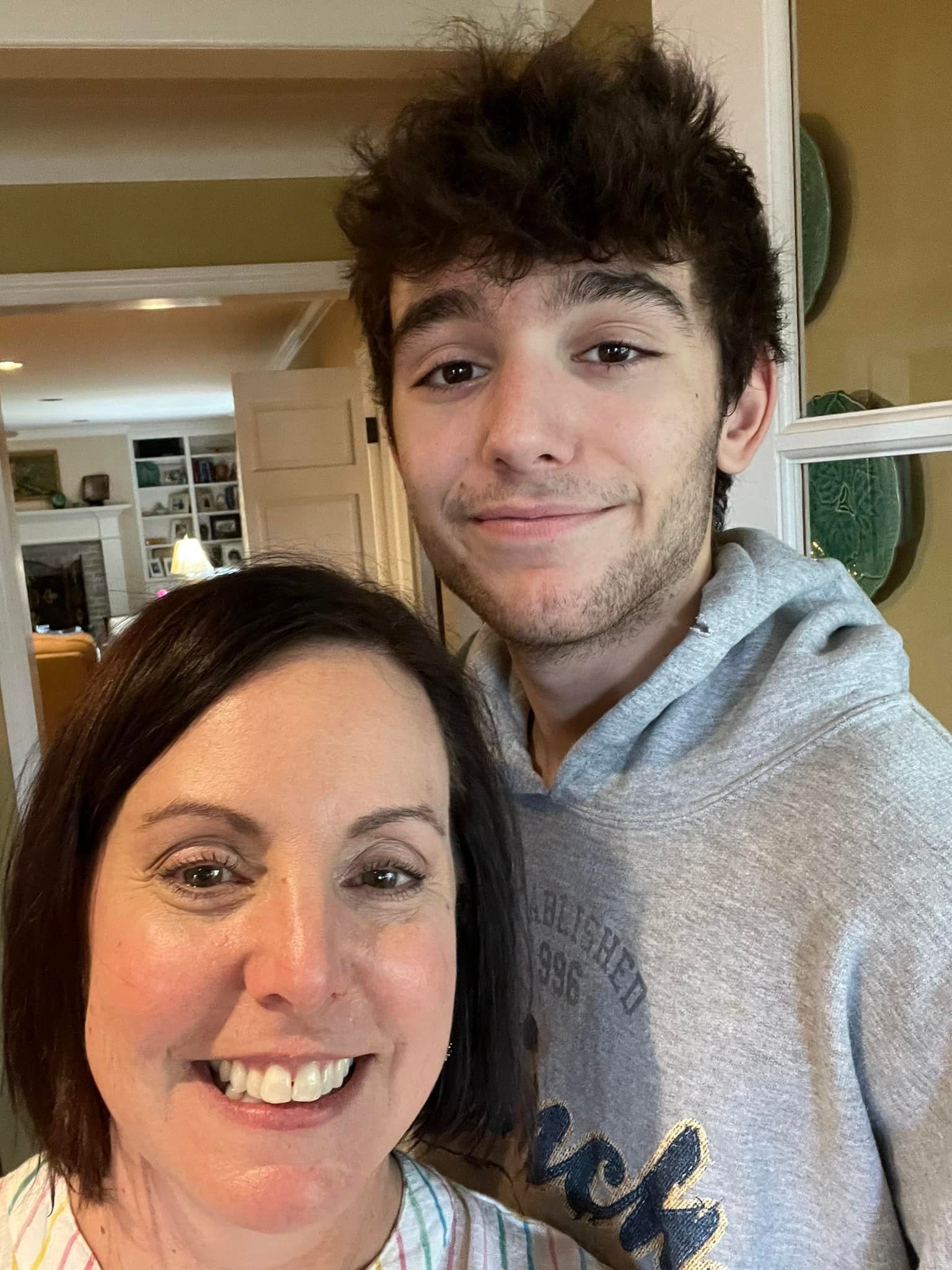 Two people smiling in a home interior, one in a gray hoodie standing behind the other.