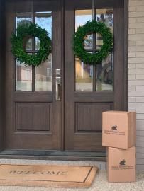 Front door with two green wreaths, a doormat, and two stacked cardboard boxes on the porch.