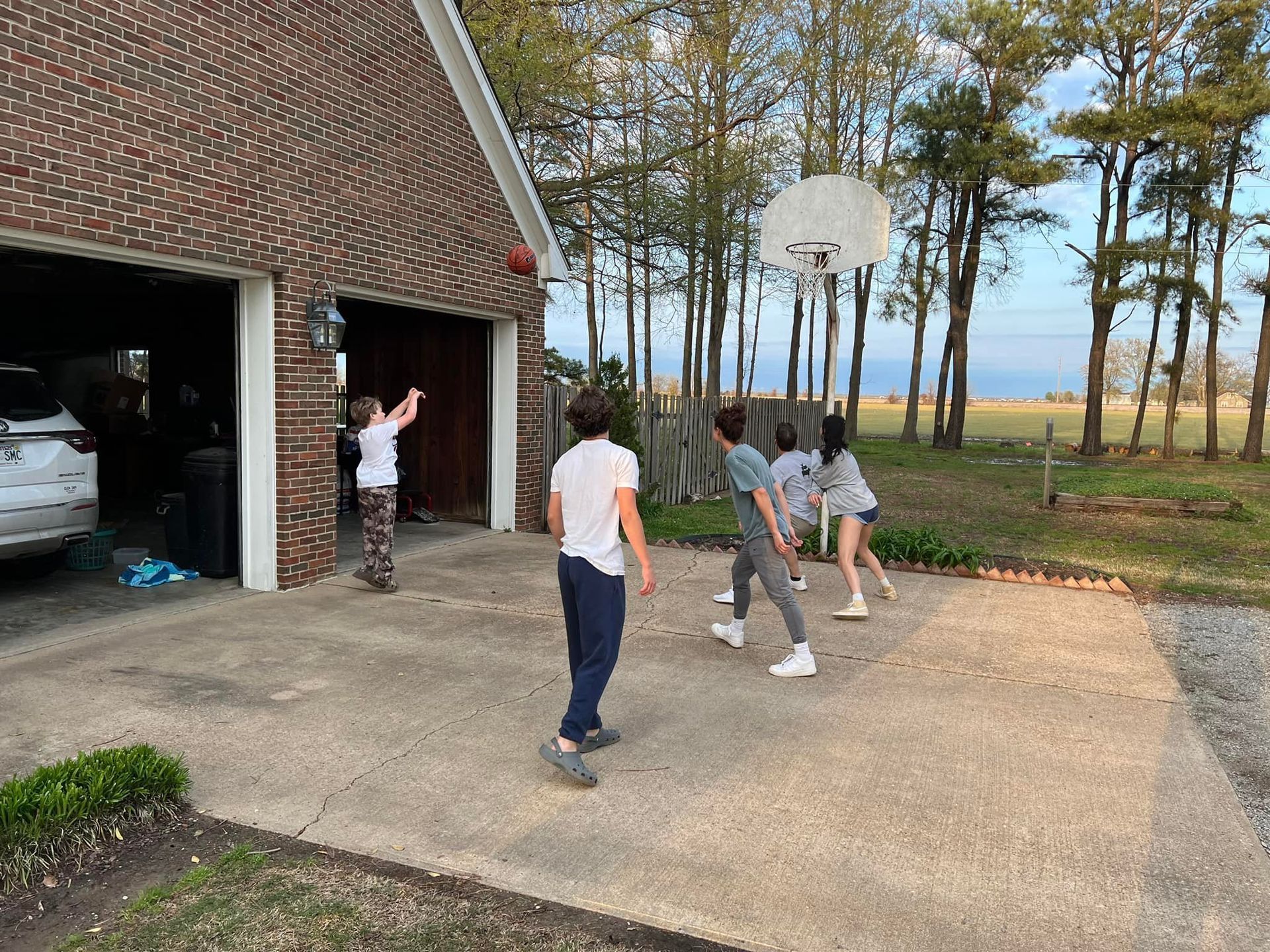 Children playing basketball on a driveway beside a brick garage, with trees and open land in the background.