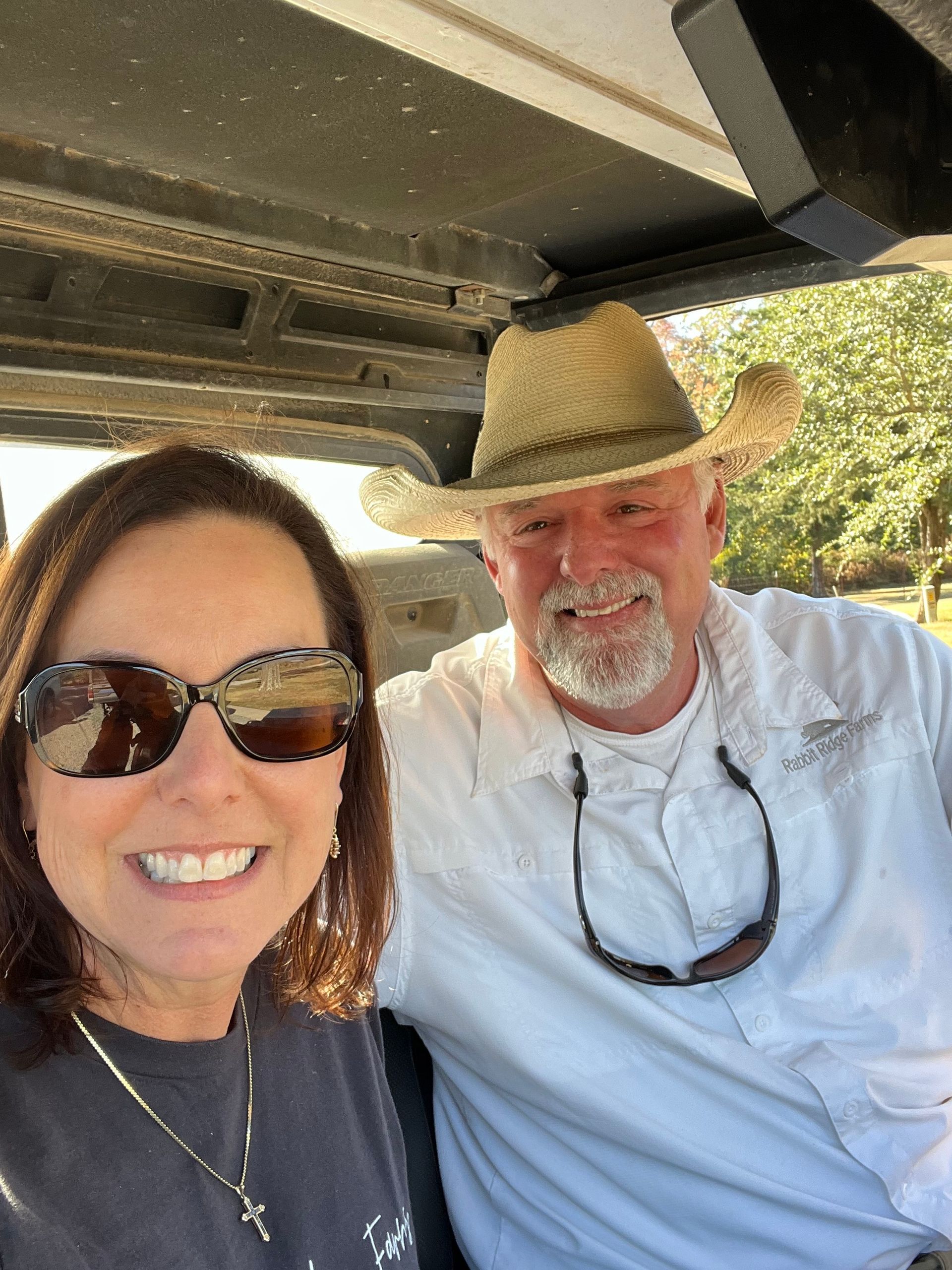 Two smiling people taking a selfie under a shaded outdoor structure, wearing sunglasses and a straw hat.