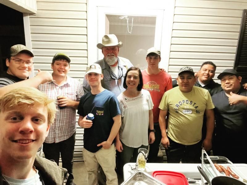 Group of smiling people gathered on a porch, holding drinks and posing for a selfie at night.