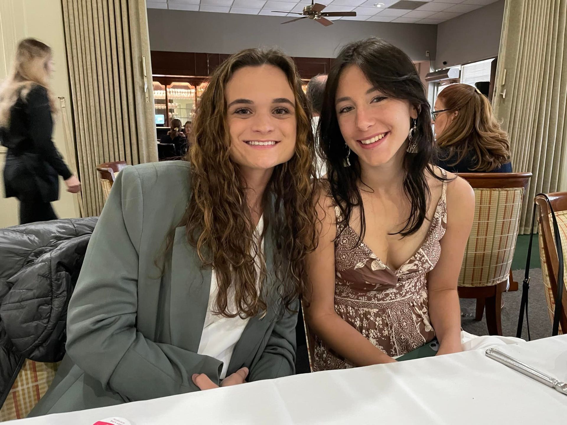 Two smiling people seated at a table in a restaurant, wearing a gray blazer and a patterned dress.