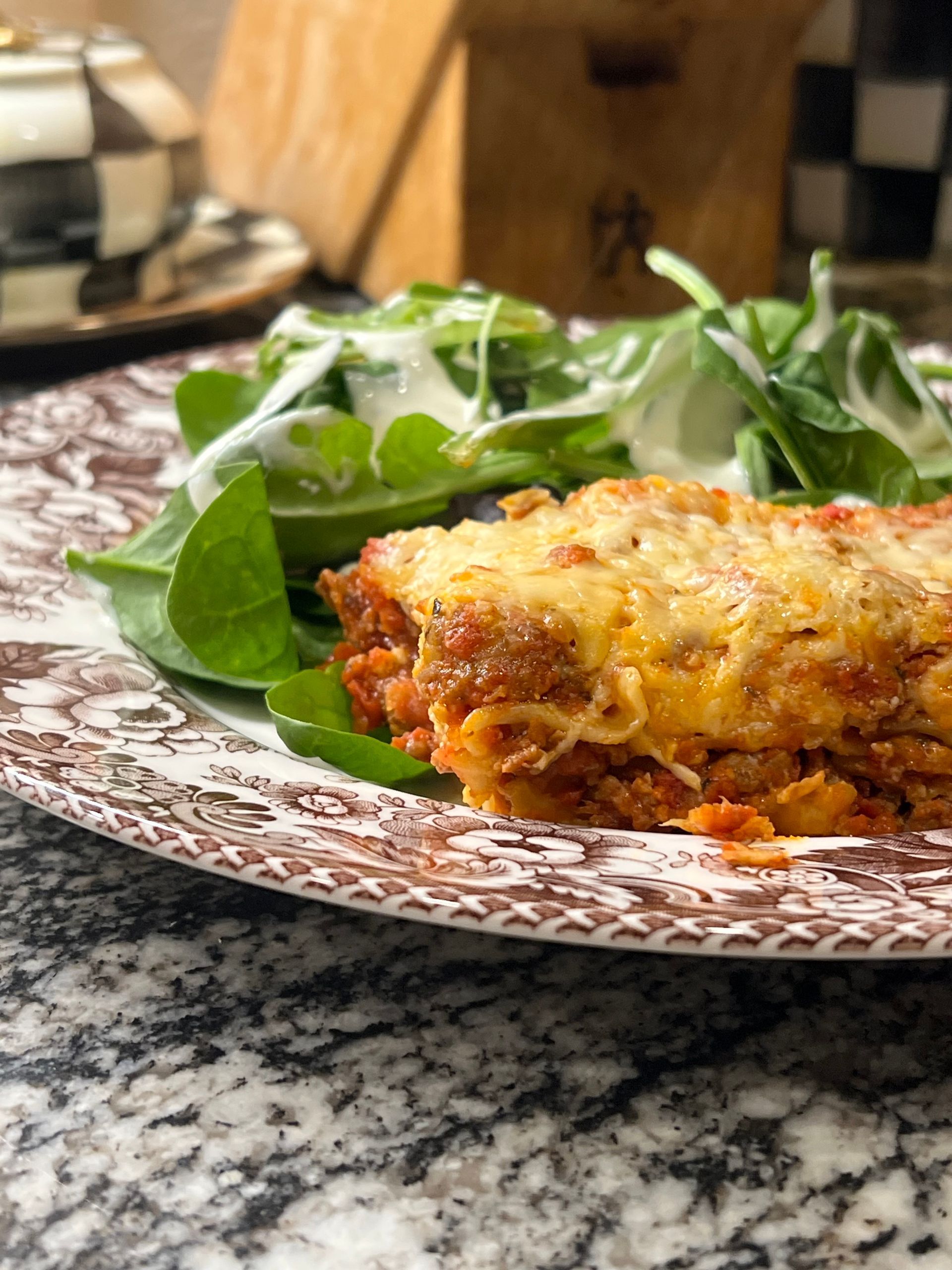 Baked lasagna with melted cheese on a patterned plate beside a green salad on a kitchen counter