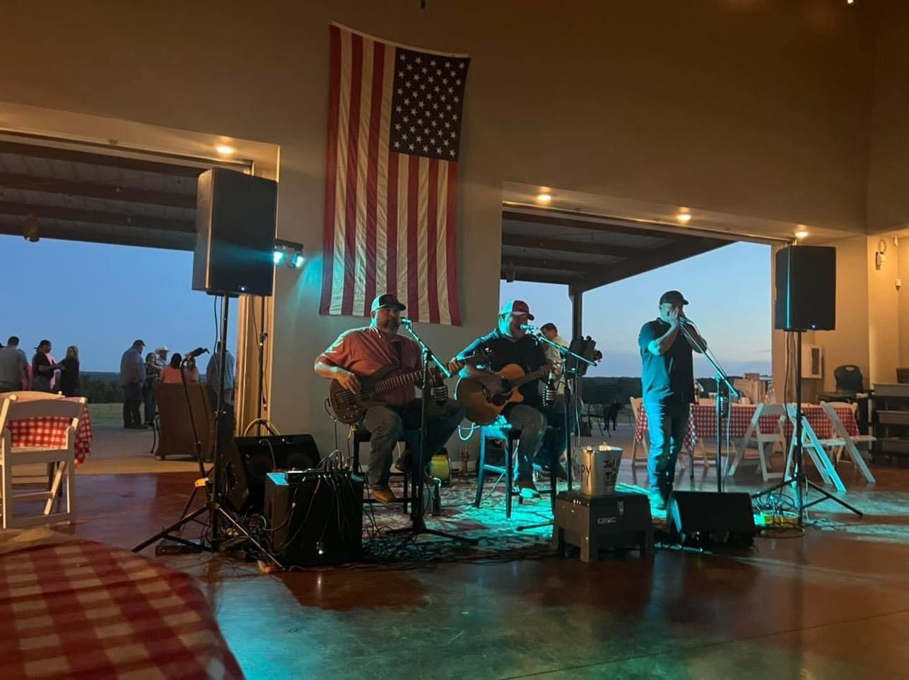 Live band performing on a patio under an American flag at dusk, with teal stage lighting.