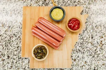 Hot dogs on a wooden board with three bowls of condiments on a granite countertop