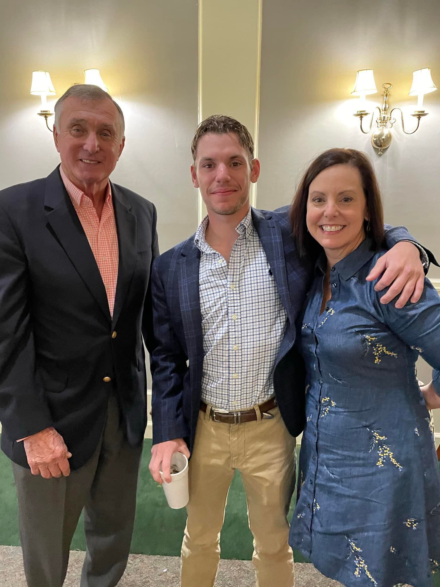 Three people smiling indoors at a formal event, with wall sconces in the background.