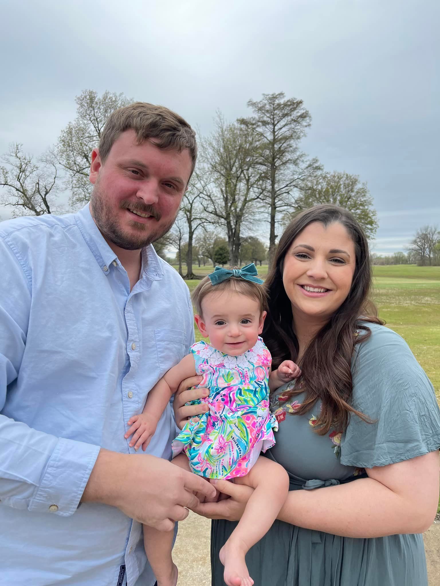 Family of three posing outdoors; man and woman hold a baby in a floral dress on a cloudy day.
