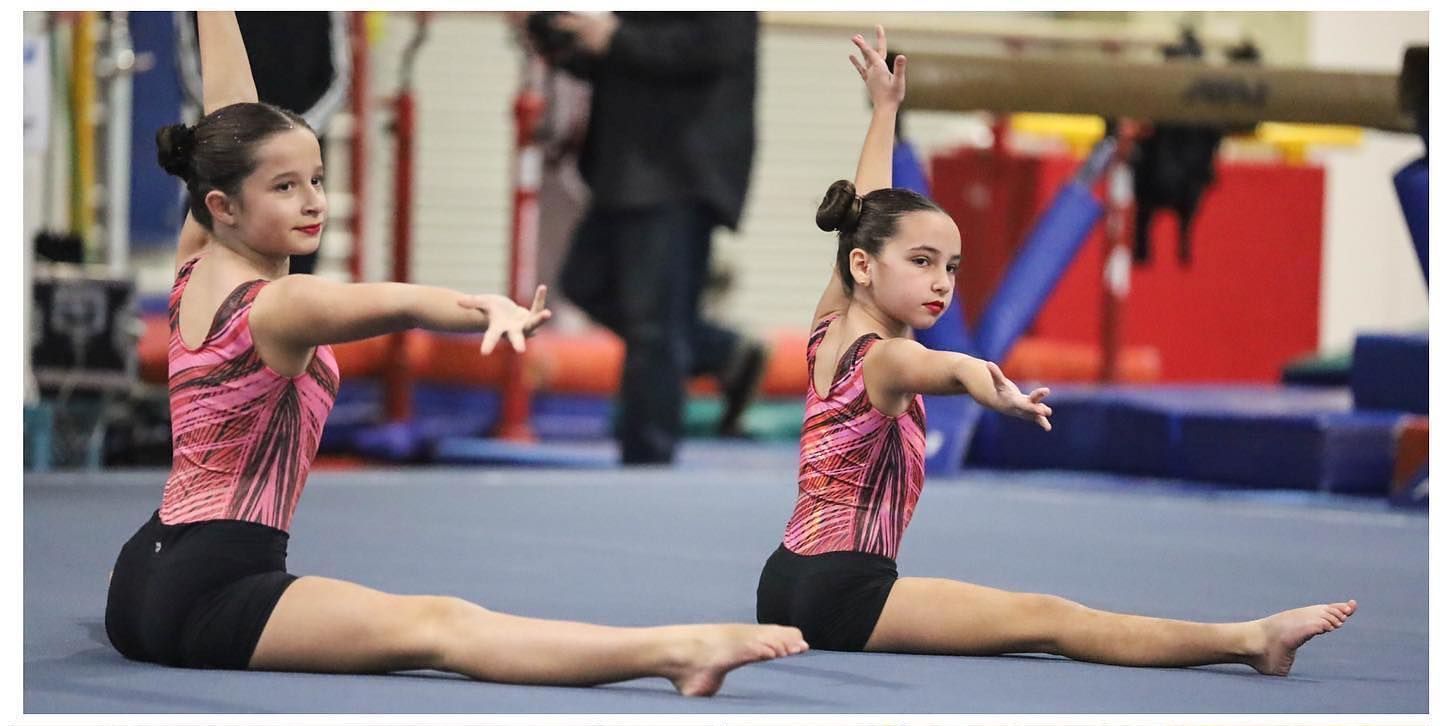 Two girls in matching leotards and shorts sitting on a gym floor performing a routine.