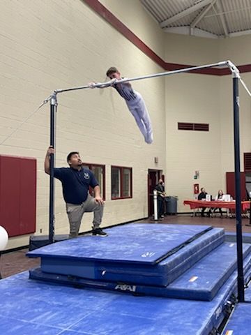 A man is watching a child do a trick on a bar in a gym.