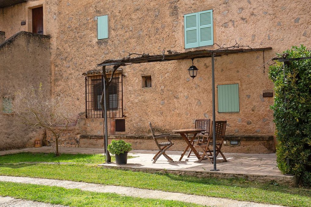 Exterior de una casa rústica con un pequeño patio y zona de estar. Paredes de estuco marrón claro y contraventanas verde pálido.