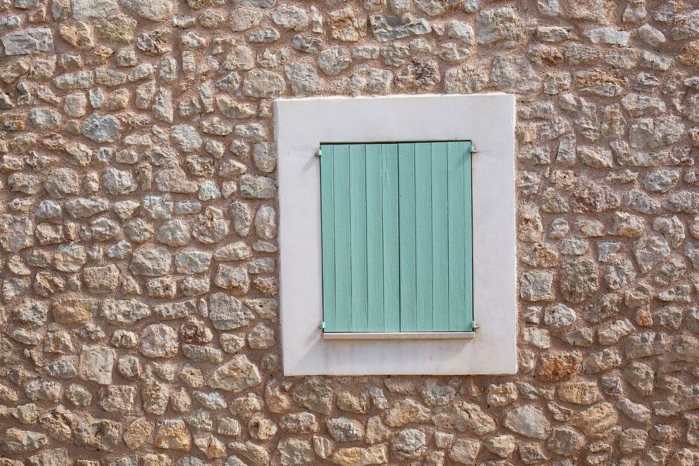 Ventana con contraventanas color verde azulado colocadas en un marco blanco, contra una pared de piedra texturizada.