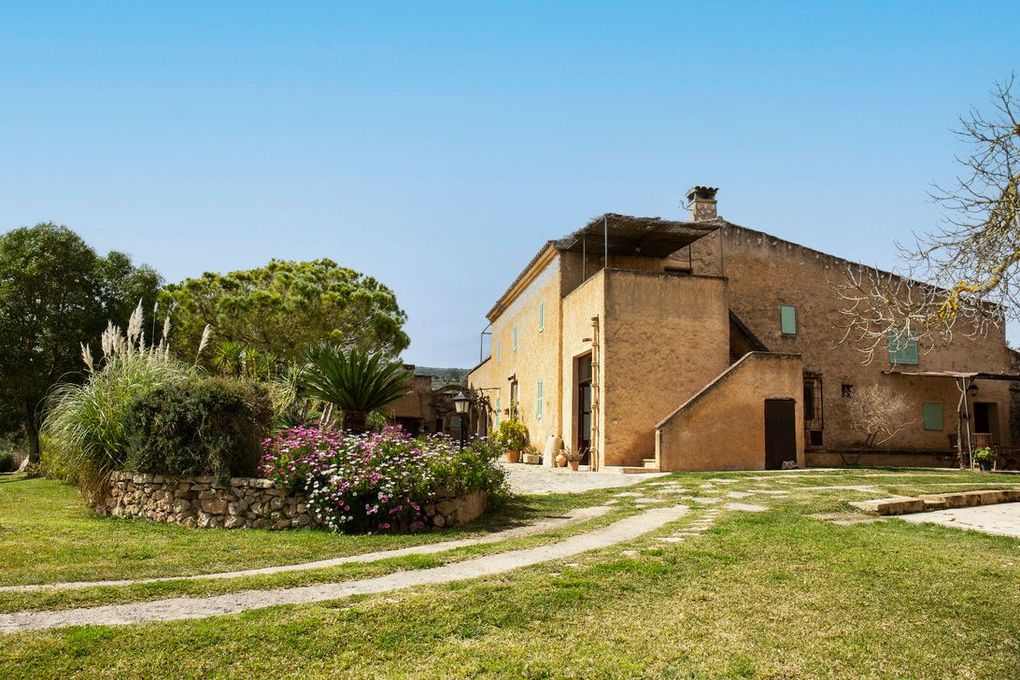 Casa de piedra con entrada de tierra, rodeada de vegetación y bajo un cielo azul claro.