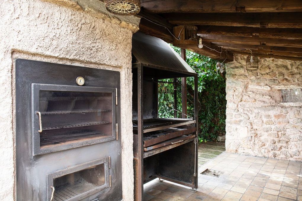 Estructura exterior de piedra con parrilla metálica y horno integrados. Zona cubierta con vigas de madera.