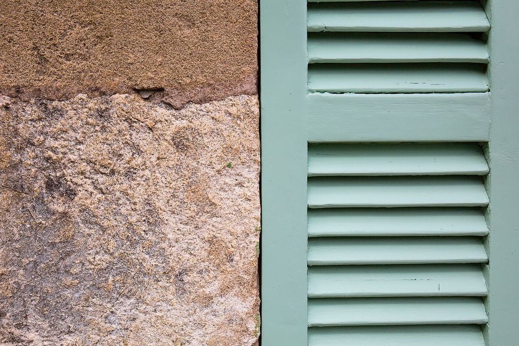Muro de piedra junto a una contraventana verde claro con lamas horizontales.