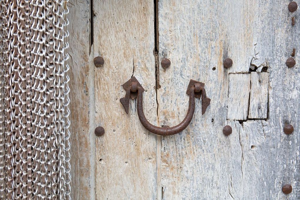 Puerta de madera vieja y desgastada, con tirador y tachuelas de metal oxidado. Una cota de malla cuelga a la izquierda.
