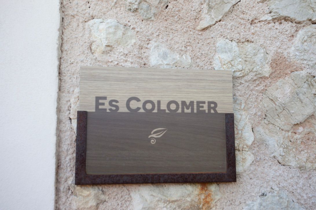 Auf einem Holzschild an einer Steinmauer steht „Es Colomer“.