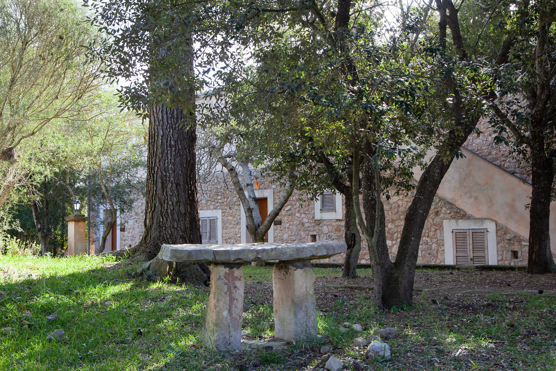 A stone table is sitting in the grass in front of a stone building surrounded by trees.