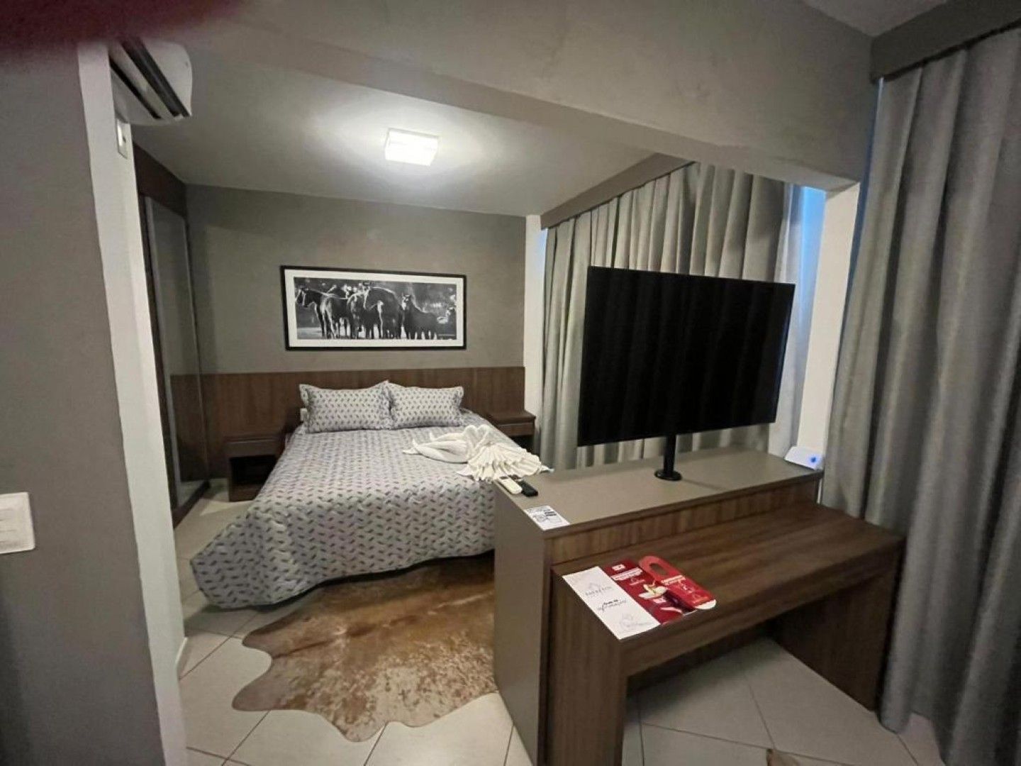 Quarto com cama, TV e escrivaninha; com tons neutros, detalhes em madeira e um tapete de couro de vaca.