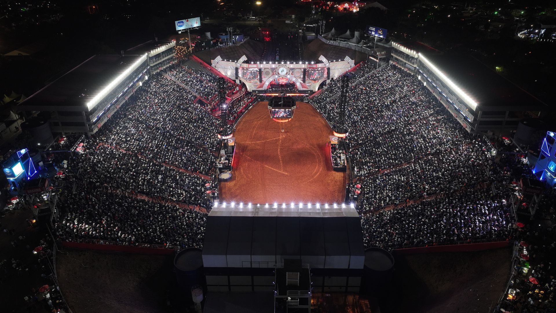 Uma arena de rodeio lotada, com piso de terra, espectadores e telões, iluminada pelo crepúsculo.
