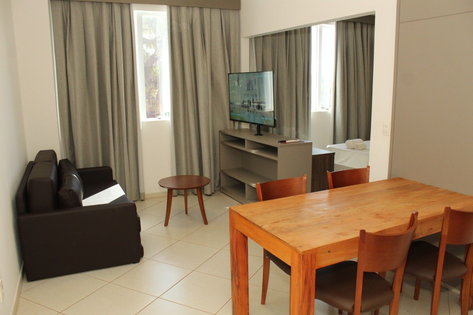 Sala de estar com sofá, mesa, cadeiras e TV; cortinas cobrem as janelas.