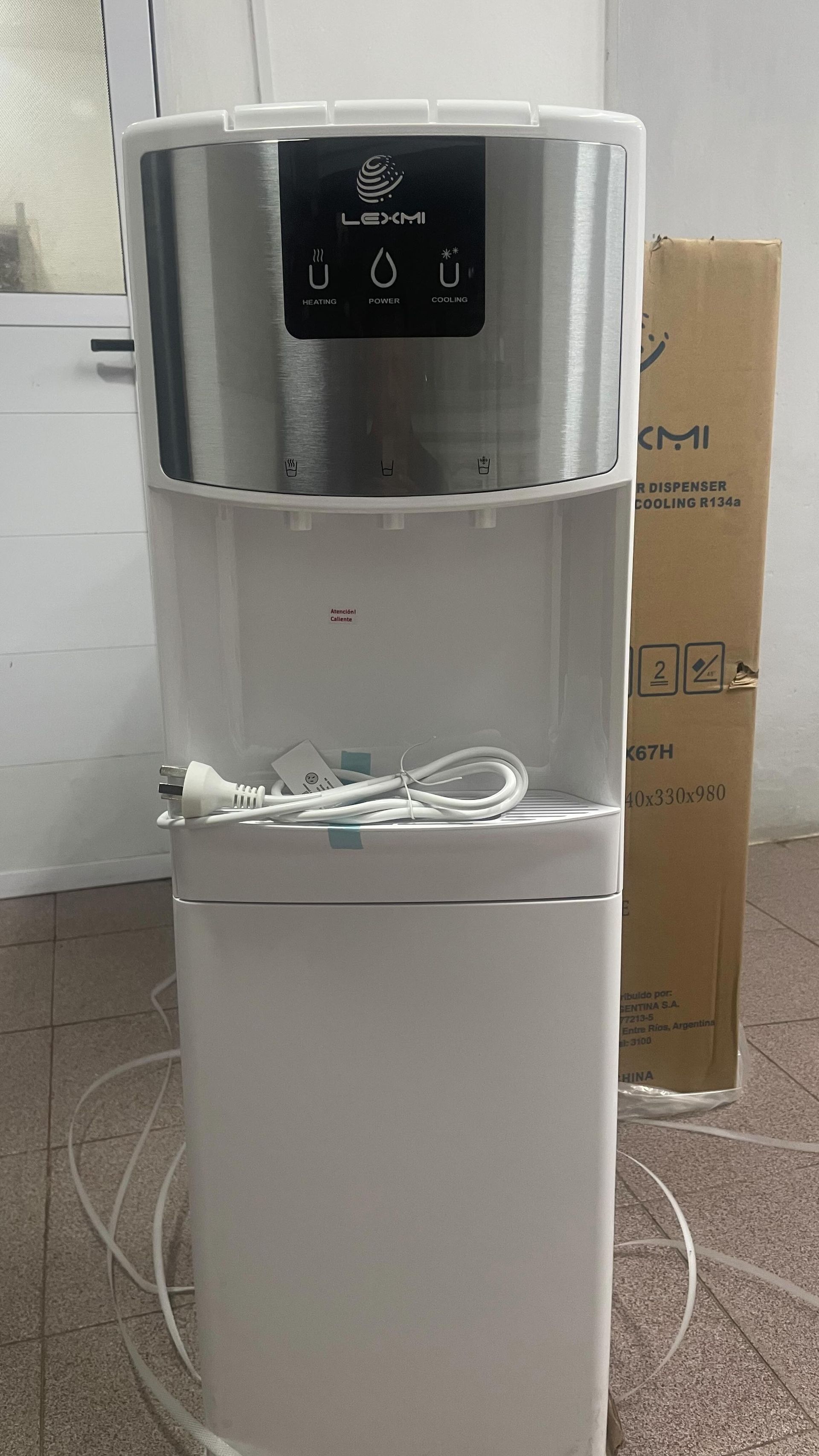 Dispensador de agua blanco con panel digital y cable en espiral en una habitación alicatada, cajas detrás.