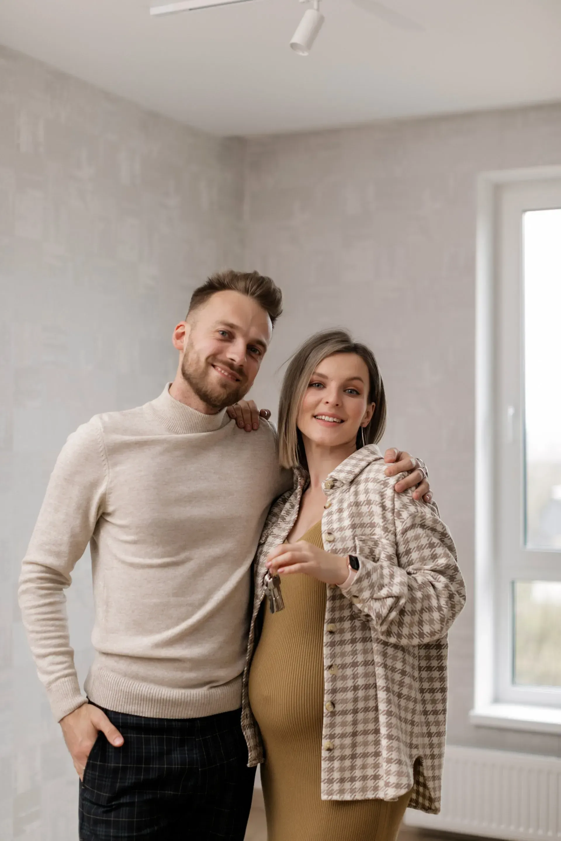 Couple posing indoors, smiling beside a window in a bright, neutral-toned room