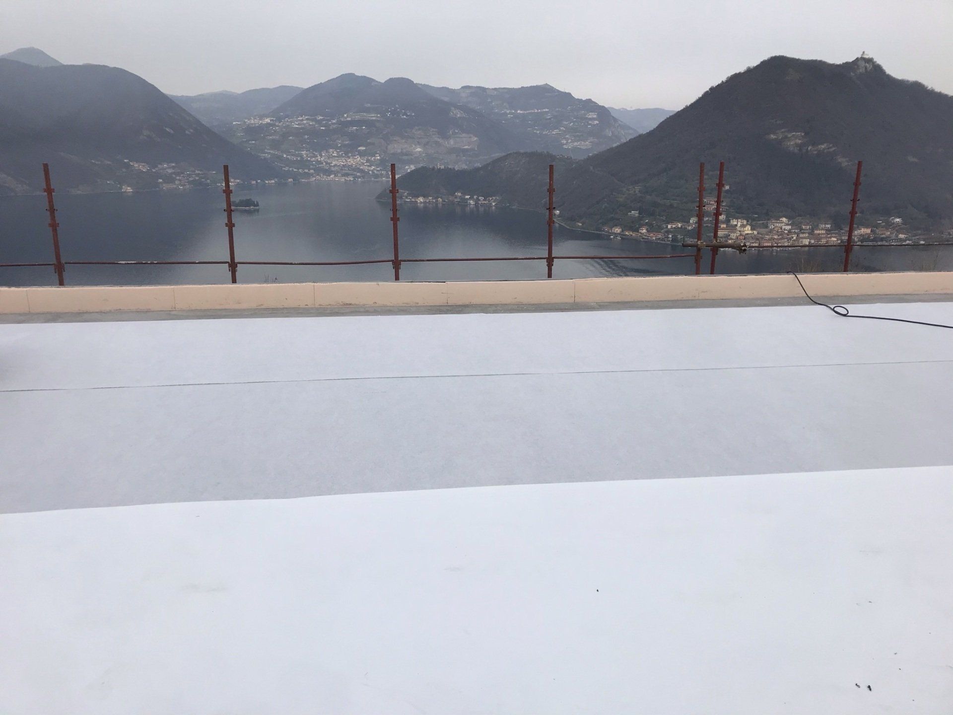 Terrazza innevata con vista su montagne avvolte nella nebbia e su un lago, con una ringhiera in primo piano.