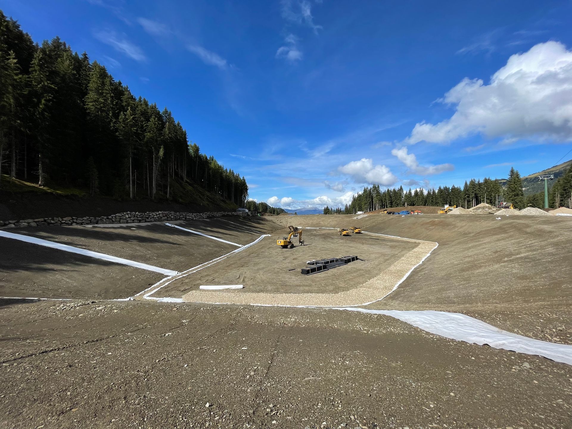 Cantiere edile in ghiaia su un pendio collinare con pineta, nastri di contenimento bianchi e un cielo azzurro con nuvole.