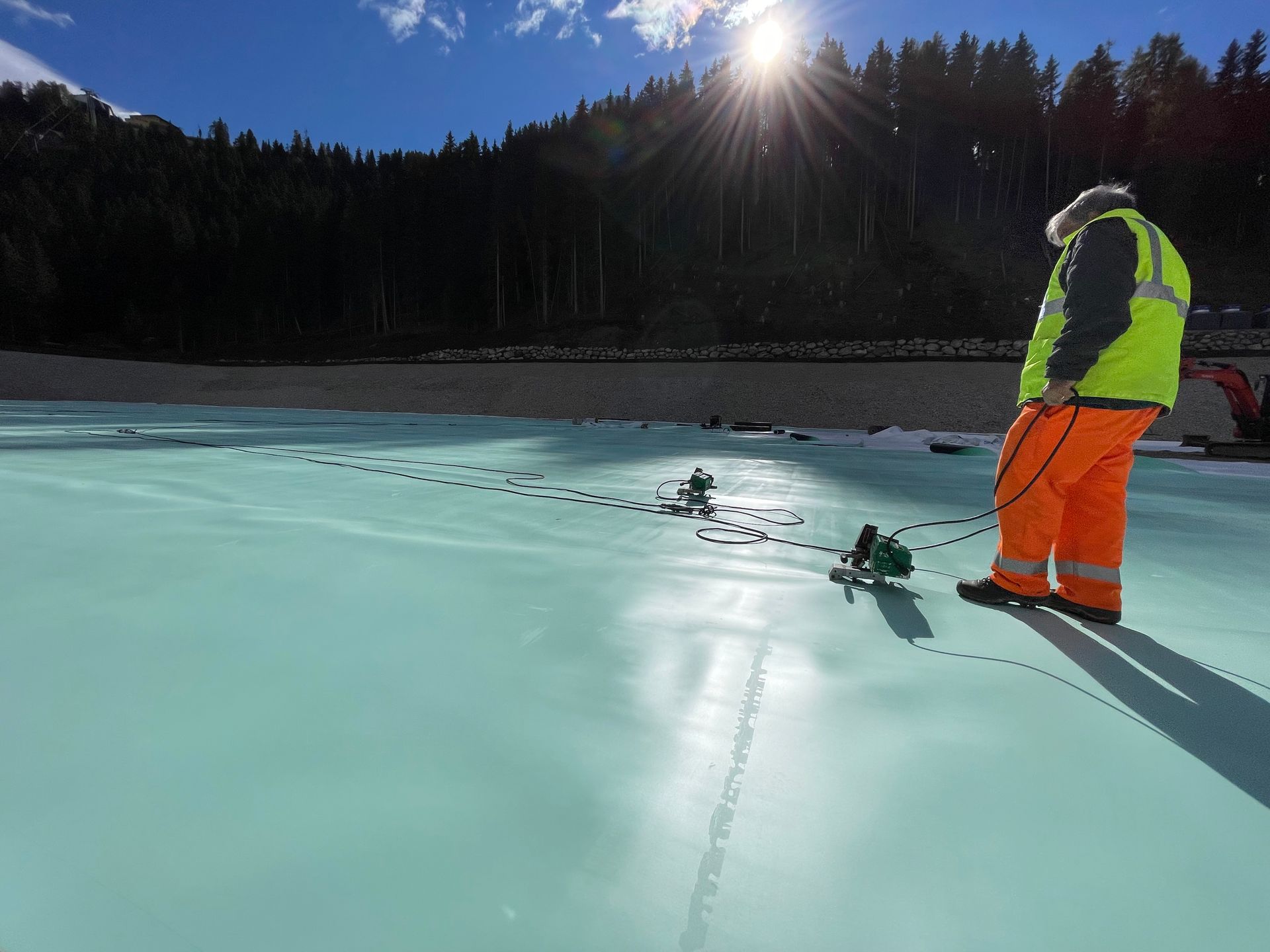 Un operaio con giacca fluorescente e pantaloni arancioni è in piedi su uno stagno ghiacciato sotto un sole splendente, con la riva boscosa sullo sfondo.