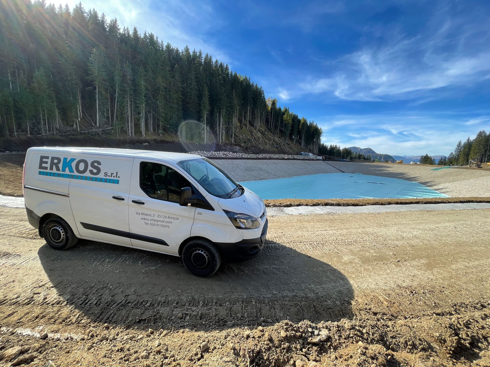 Furgone di servizio bianco della ERKOS parcheggiato su una riva ghiaiosa accanto a un lago blu e a una pineta.
