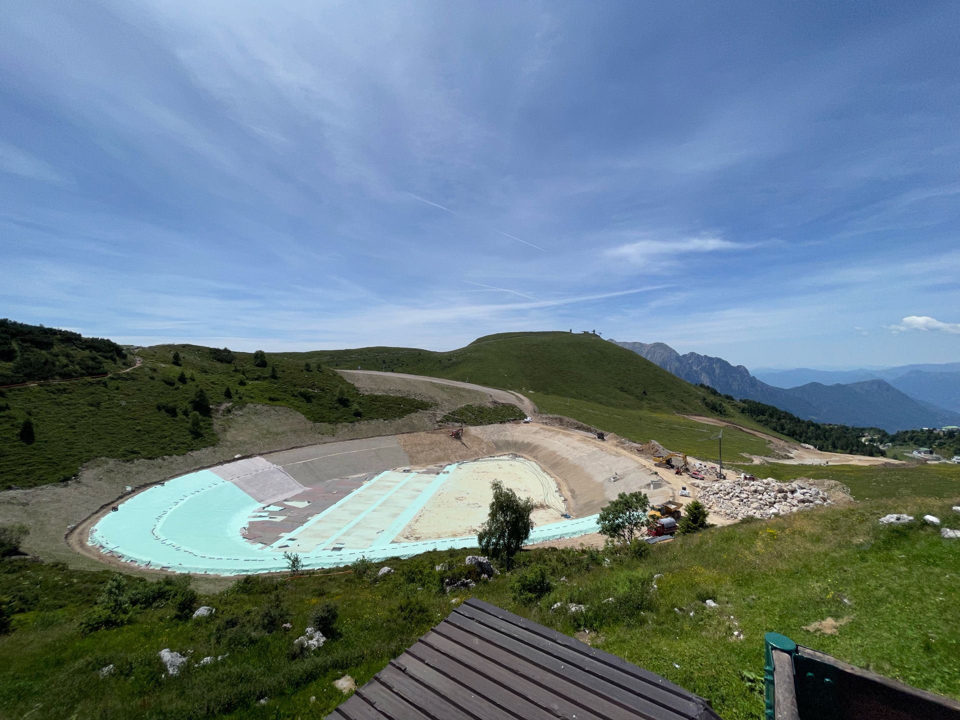 Pendio montuoso con bacino di costruzione di colore azzurro pallido, strade sterrate e lontane cime blu sotto un cielo limpido.