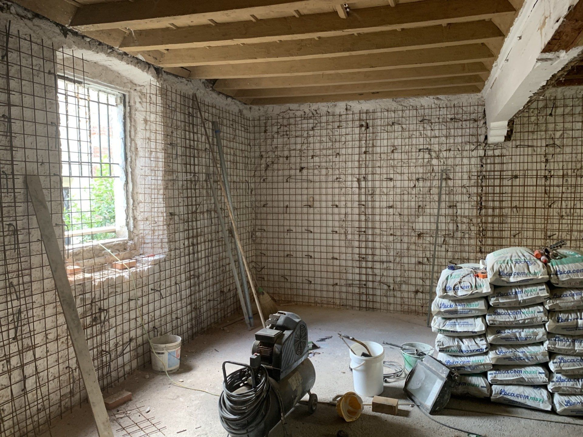Stanza non rifinita con pareti in mattoni a vista, travi a vista sul soffitto, una finestra e materiali edili sul pavimento.