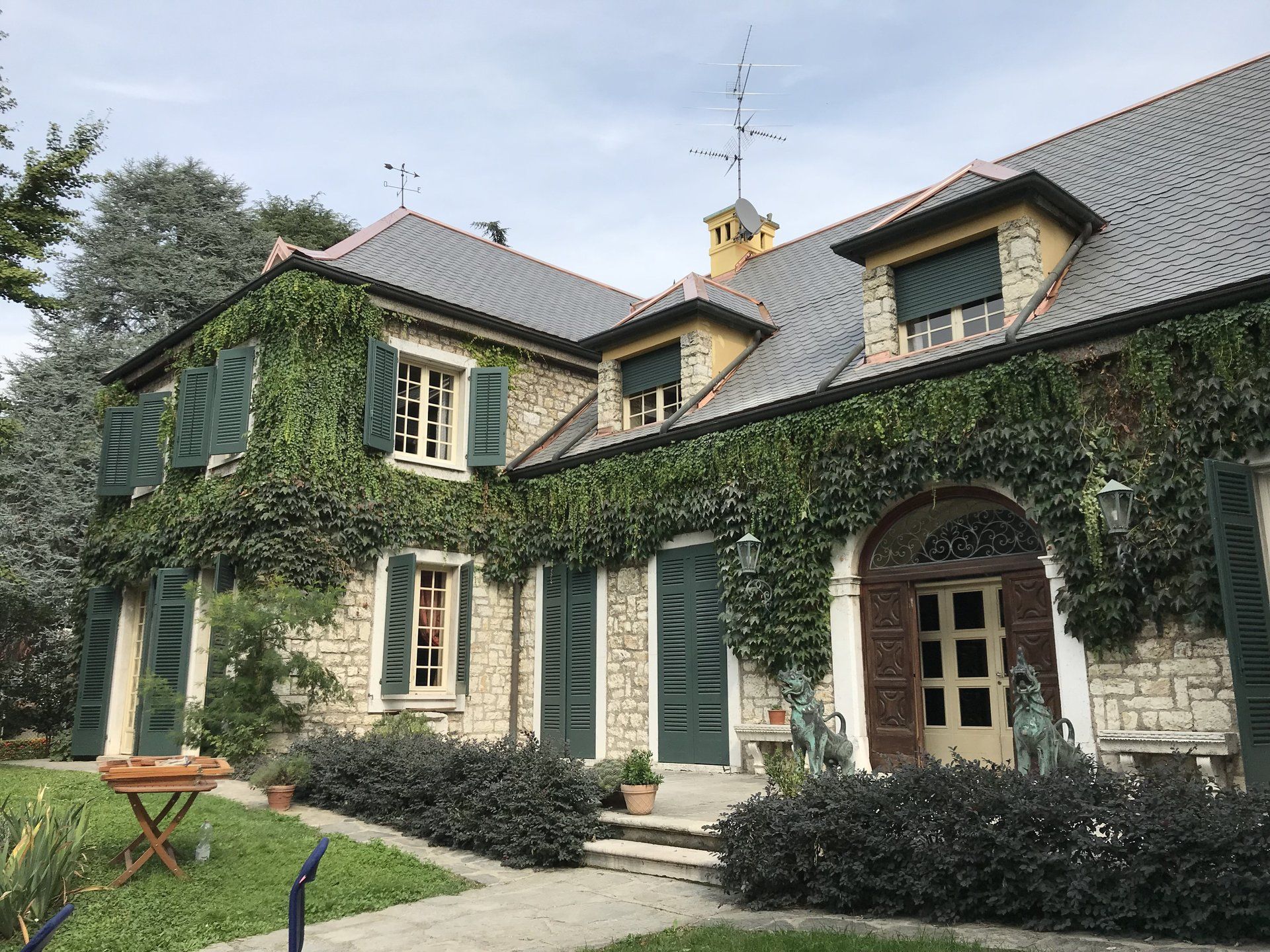 Casa in pietra ricoperta di edera con persiane verdi e tetto di tegole in un cortile con giardino.