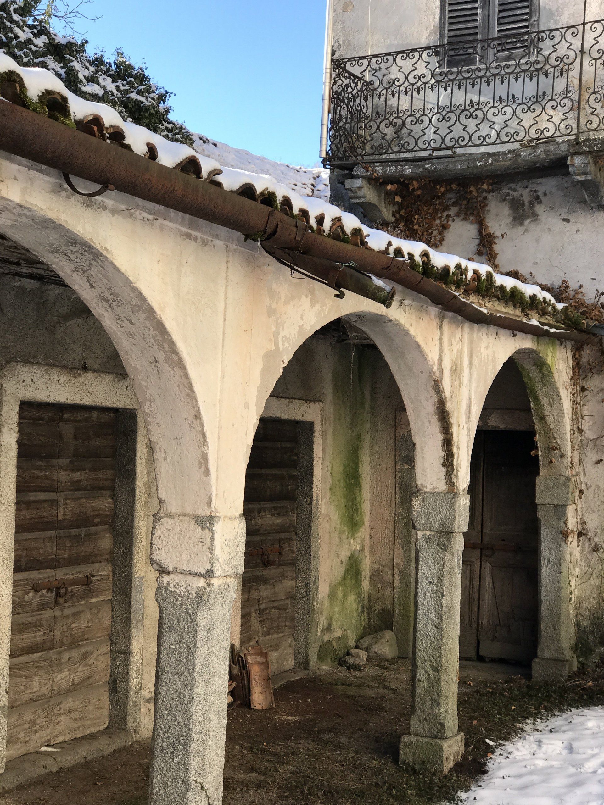 Antico cortile in pietra con colonnato ad archi, muri consumati dal tempo e un balcone in ferro battuto sovrastante.