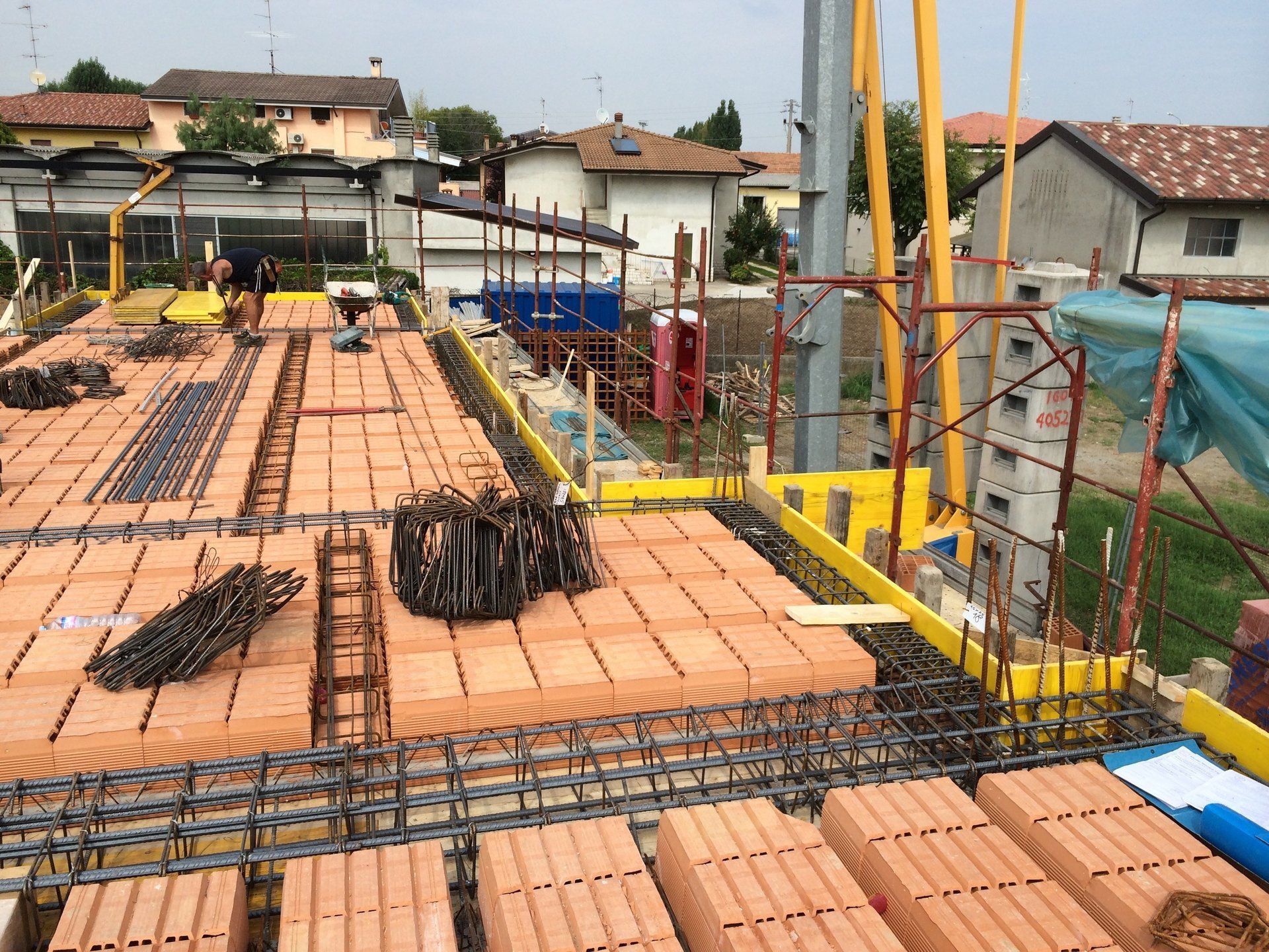 Cantiere edile con blocchi di mattoni per il pavimento, barre d'armatura, impalcature e operai sullo sfondo.